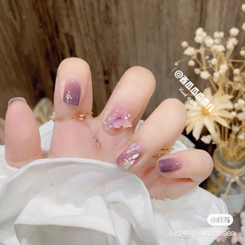 Bàn tay với móng tay màu tím trang trí hoa và ánh kim, đeo vòng tay mảnh. Phông nền có hoa khô làm điểm nhấn.