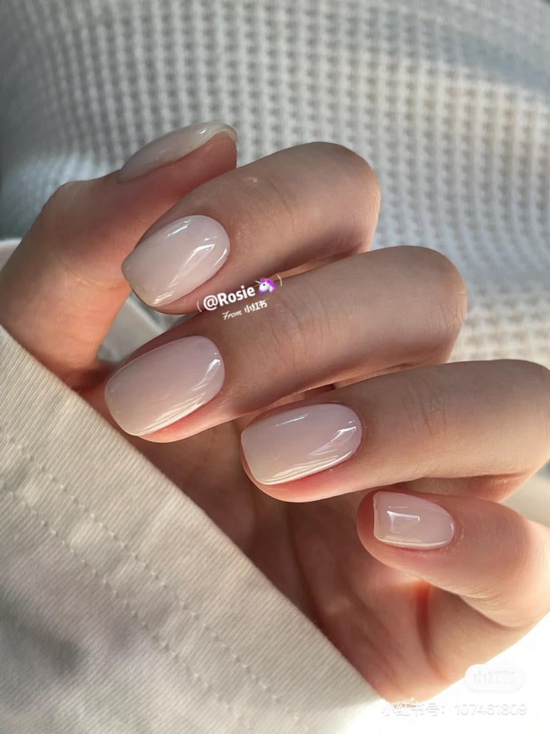 Một bàn tay với móng tay được sơn màu nude bóng, trên nền vải trắng.