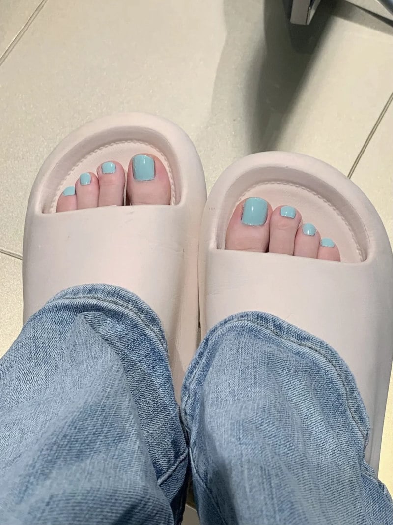 Người mang dép trắng, móng chân sơn màu xanh lam, mặc quần jeans xanh nhạt, đứng trên nền nhà lát gạch.