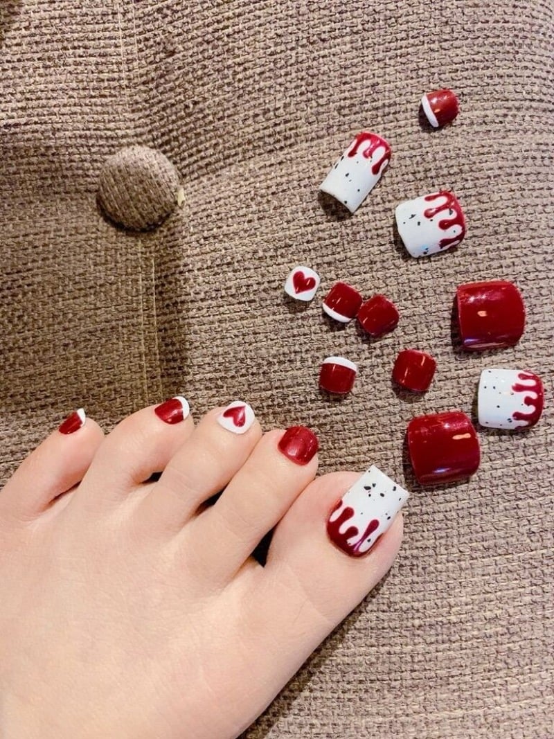 mau-nail-chan-noel__1__016b5c4afa444ef08998dd786da0cd08 Móng chân được sơn màu đỏ và trắng với họa tiết trái tim và sọc, nằm trên bề mặt vải nâu.