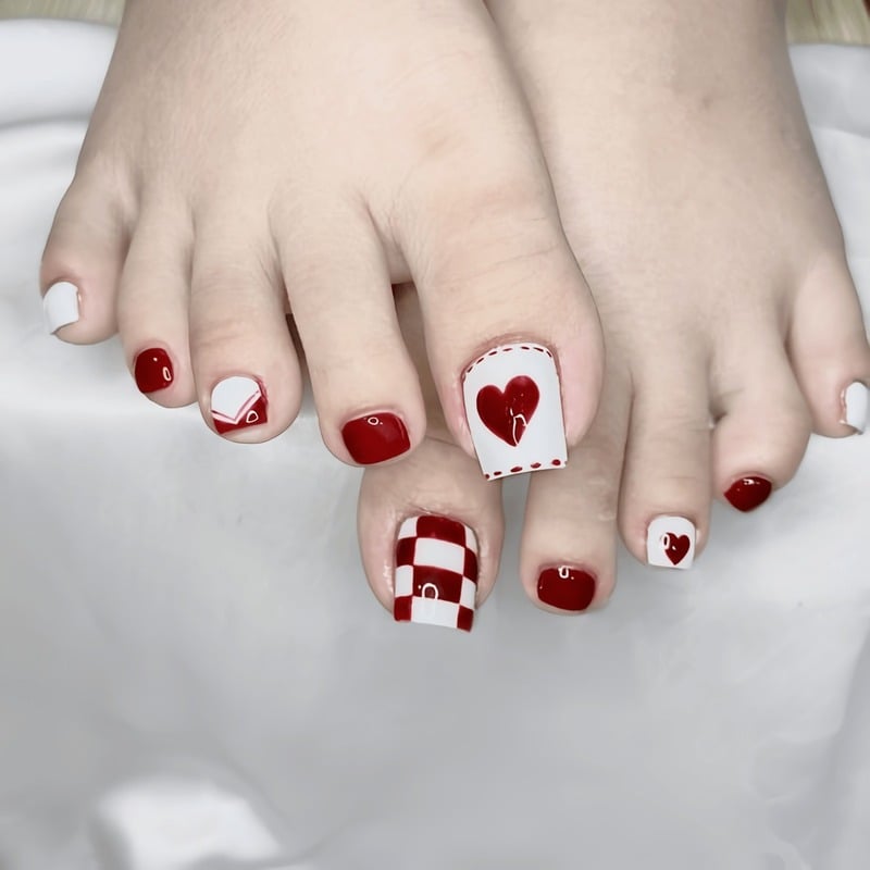 mau-nail-chan-noel__4__96b66104da424ea6b78549be246024a9 Bàn chân với móng được sơn màu đỏ và trắng, họa tiết trái tim và ô vuông trên nền vải trắng.