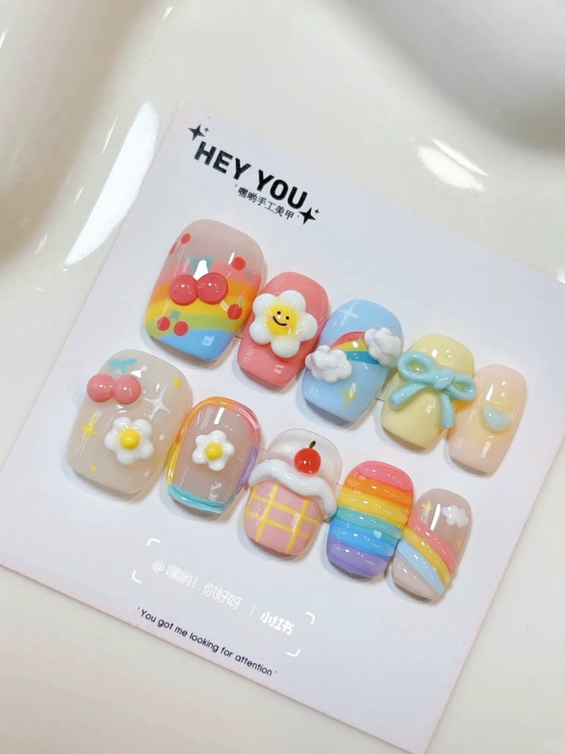 Mẫu móng tay giả với họa tiết hoa, cầu vồng và nơ, nhiều màu sắc rực rỡ trên nền trắng.