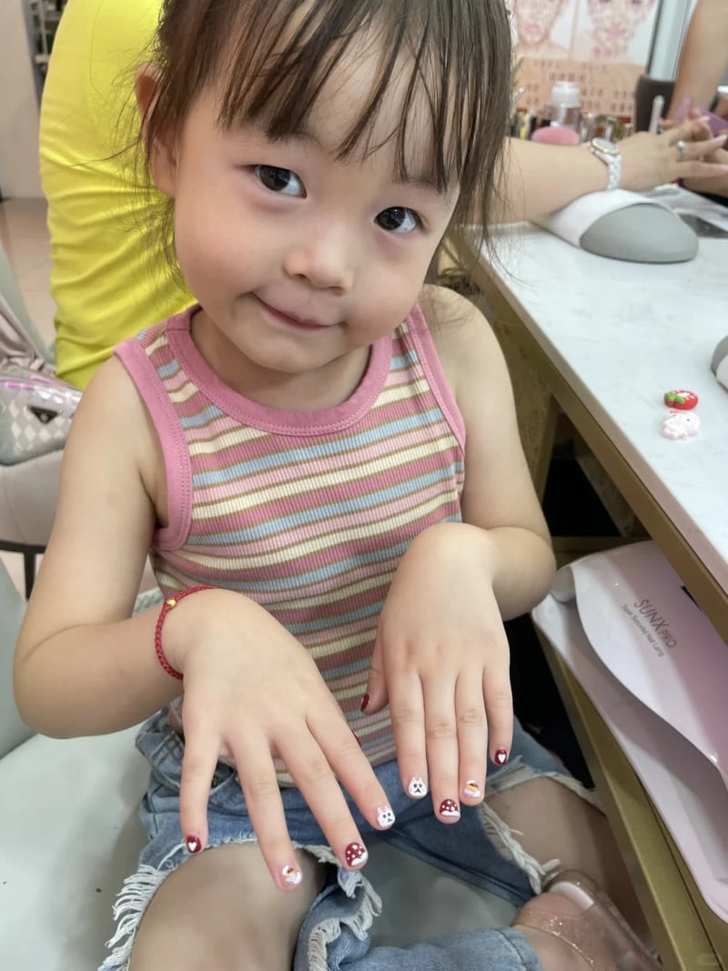 Em bé đang ngồi, giơ hai tay với móng tay được trang trí họa tiết nấm, mặc áo sọc ngang.
