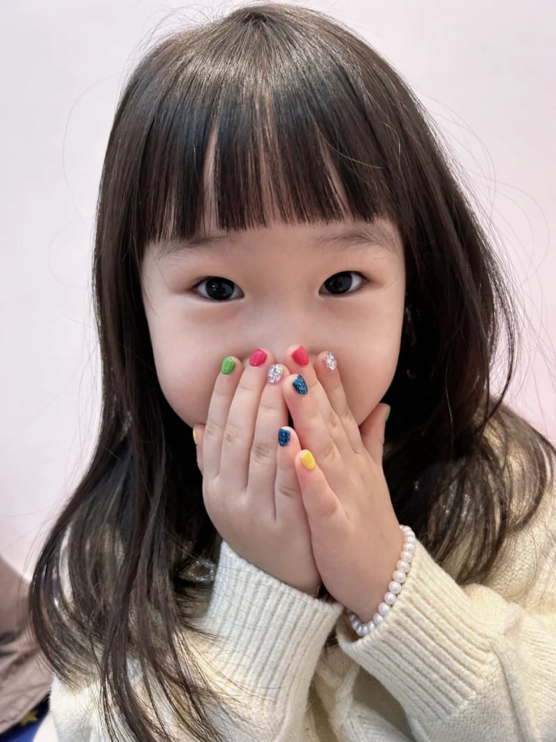 Bé gái cười, che miệng bằng hai tay, móng tay sơn nhiều màu sắc, đeo vòng tay ngọc trai, mặc áo len trắng.