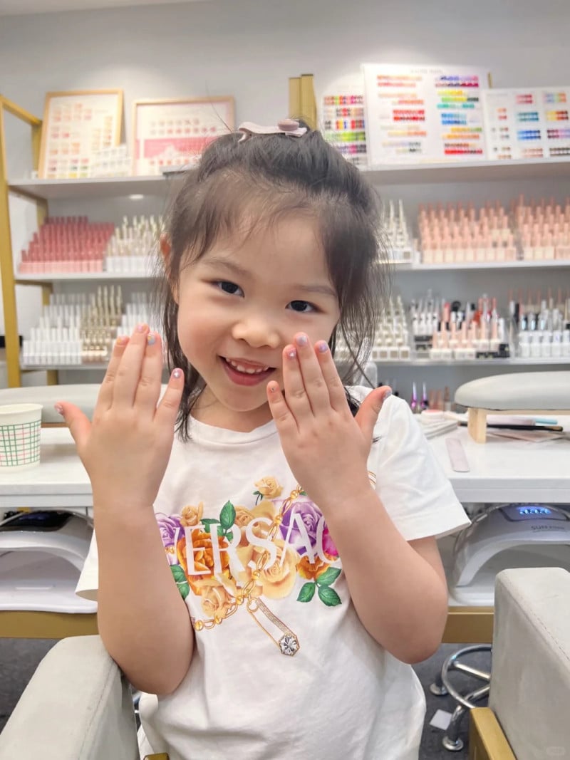 Một bé gái đang cười, giơ tay khoe móng tay mới sơn, xung quanh là các lọ sơn móng tay nhiều màu sắc.