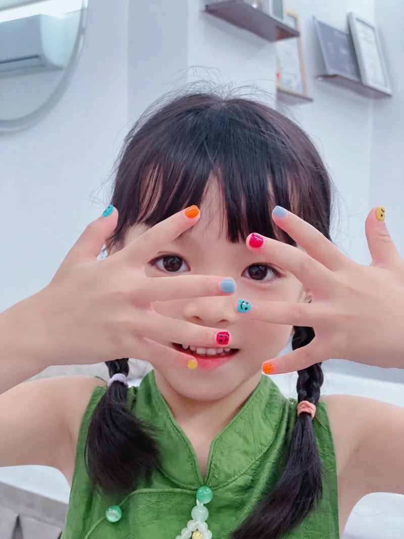 Em bé cười, tóc buộc hai bên, giơ tay với móng tay sơn màu sắc khác nhau.