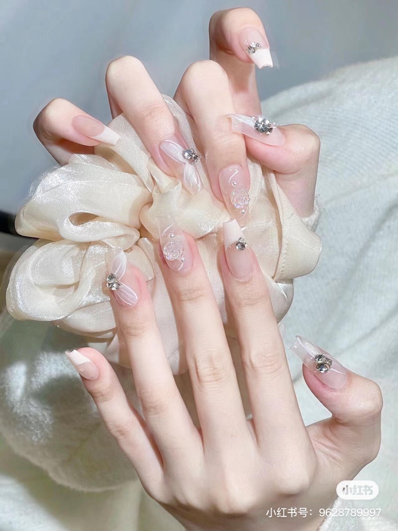 Bàn tay với móng tay dài, trang trí họa tiết 3D và đính đá, cầm một chiếc khăn voan màu be.