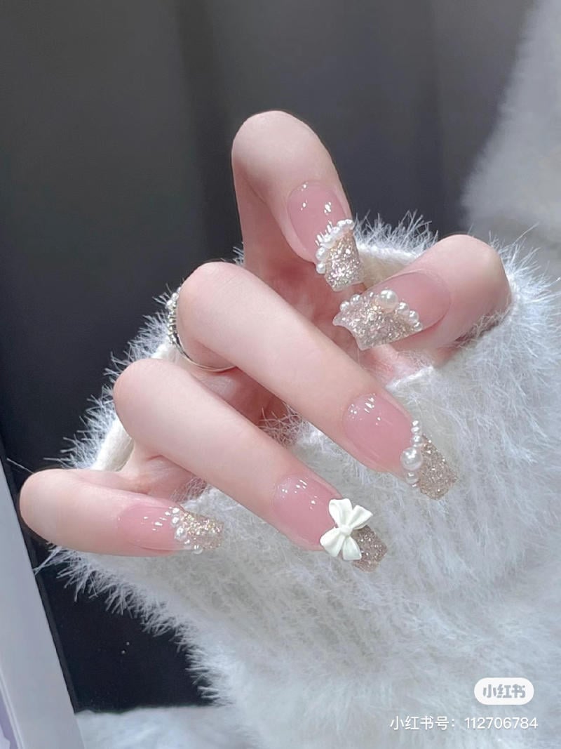 Một bàn tay với móng tay được trang trí bằng kim tuyến, ngọc trai và nơ trắng, nền áo len trắng.