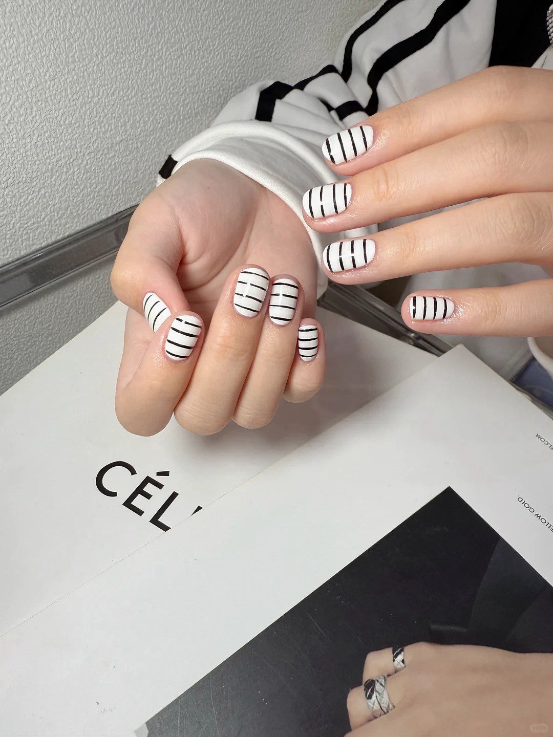 Bàn tay đeo nhẫn với móng tay sơn trắng có họa tiết kẻ đen, đặt trên mặt bàn với tạp chí.