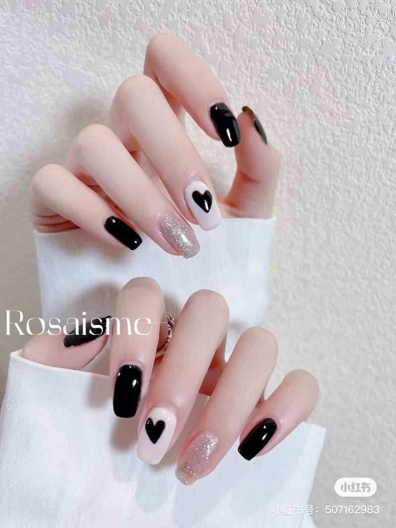 Bàn tay với móng tay sơn màu đen, trắng, nhũ bạc và họa tiết trái tim, trên nền tường trắng.