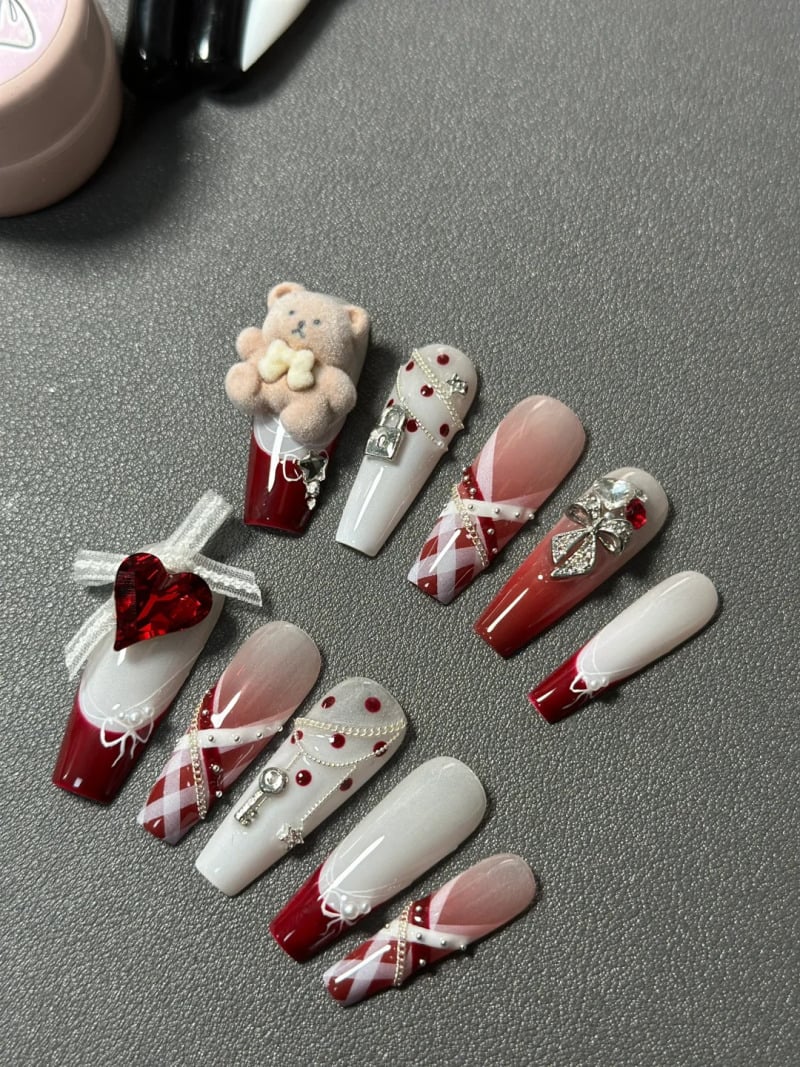 mau-nail-do-trang-cardina-17-jpg_d89da8bda8234e6697429b3049bd1e90 Mẫu móng tay giả với thiết kế đa dạng, kết hợp màu đỏ và trắng, trang trí hình gấu bông, trái tim và phụ kiện lấp lánh.