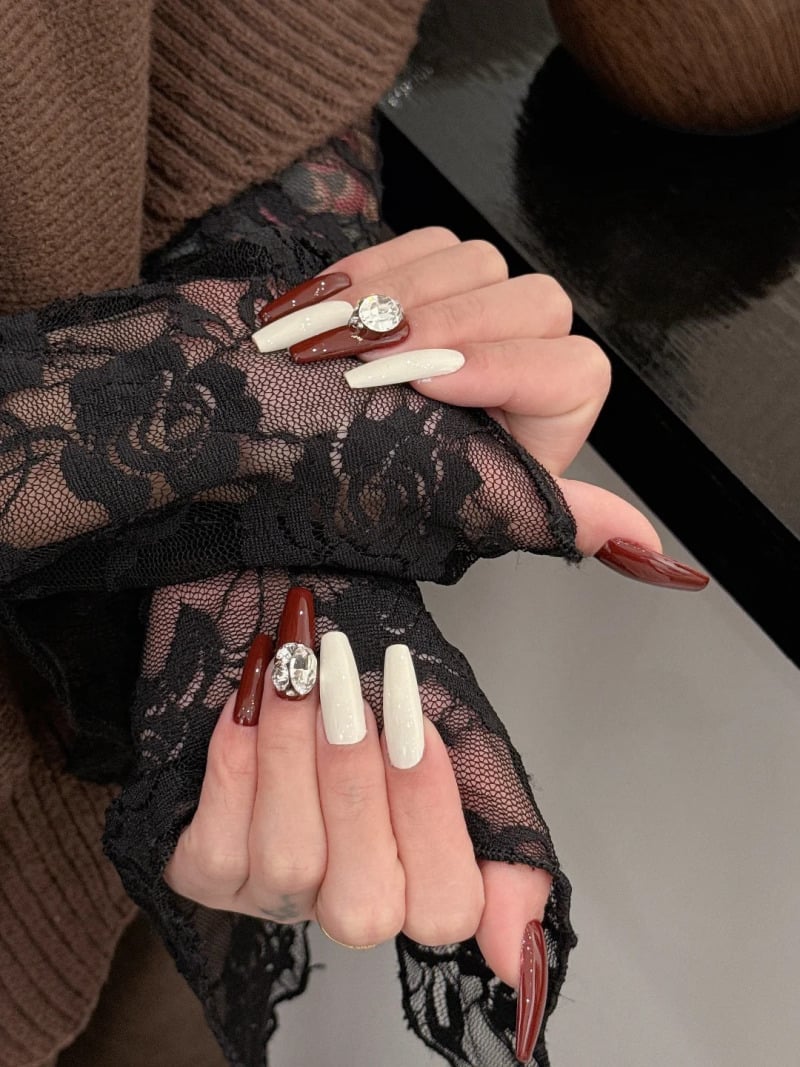 mau-nail-do-trang-cardina-19-jpg_e4aafda6e94a4bad96273d40c906413c Bàn tay với móng sơn màu đỏ và trắng, đính đá lớn, trên nền áo ren đen và áo len nâu.