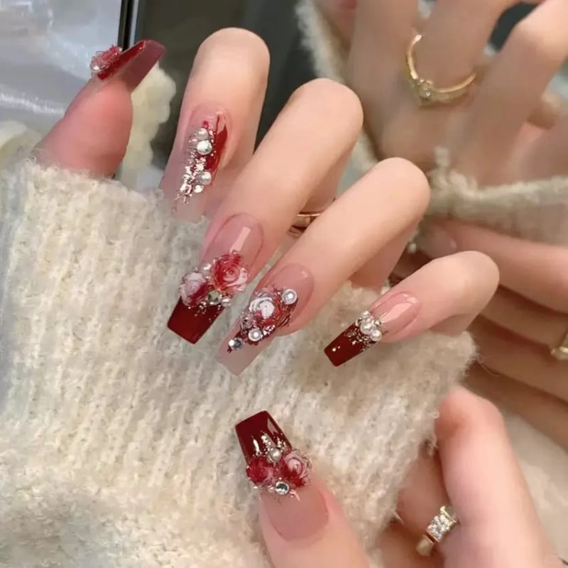 mau-nail-do-trang-cardina-21-jpg_2ab65b4d9ec54e44948f4abdc422a4ff Bàn tay với móng giả màu đỏ, họa tiết hoa và đính đá, đeo nhẫn vàng, áo len màu sáng.