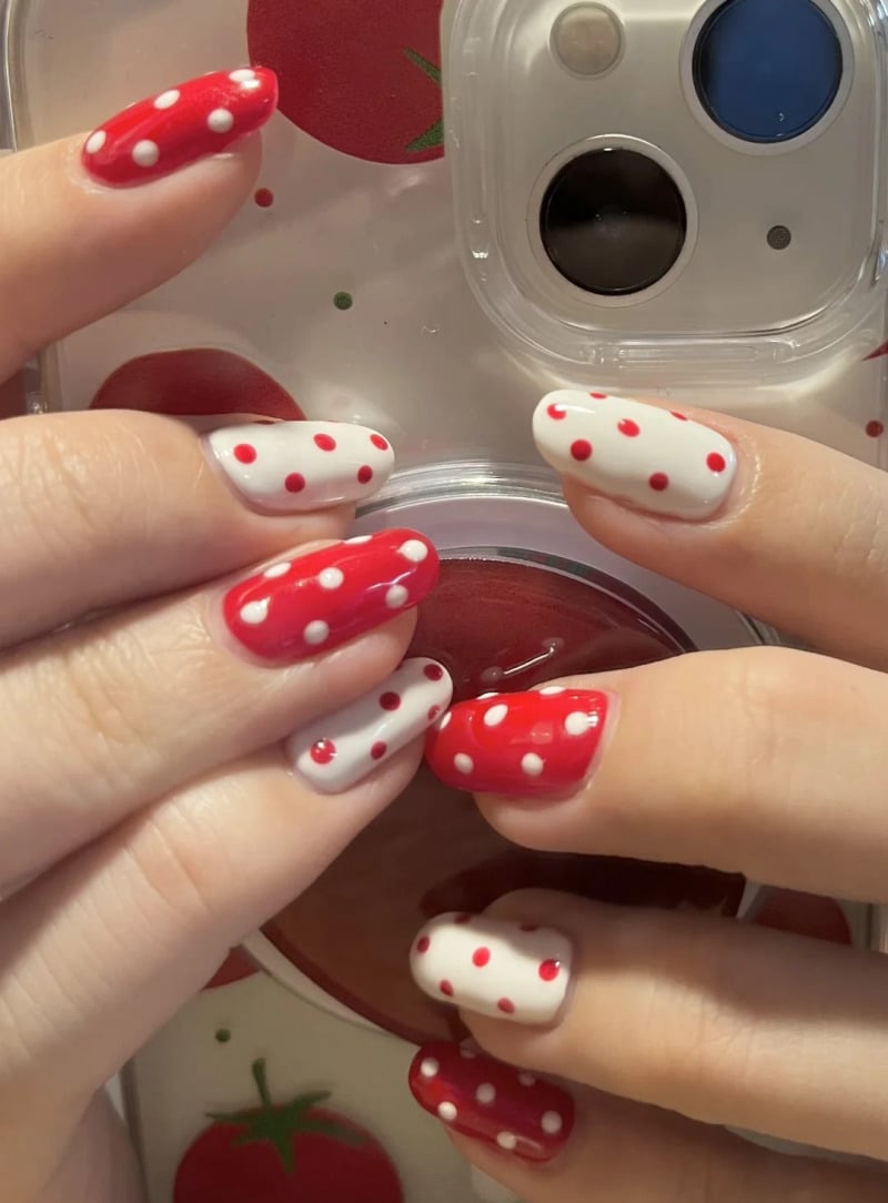 mau-nail-do-trang-cardina-3-jpg_aa1c1cf545aa413d89398b84f303276a Bàn tay với móng tay sơn đỏ và trắng chấm bi, cầm ốp lưng điện thoại trong suốt có hình quả cà chua.