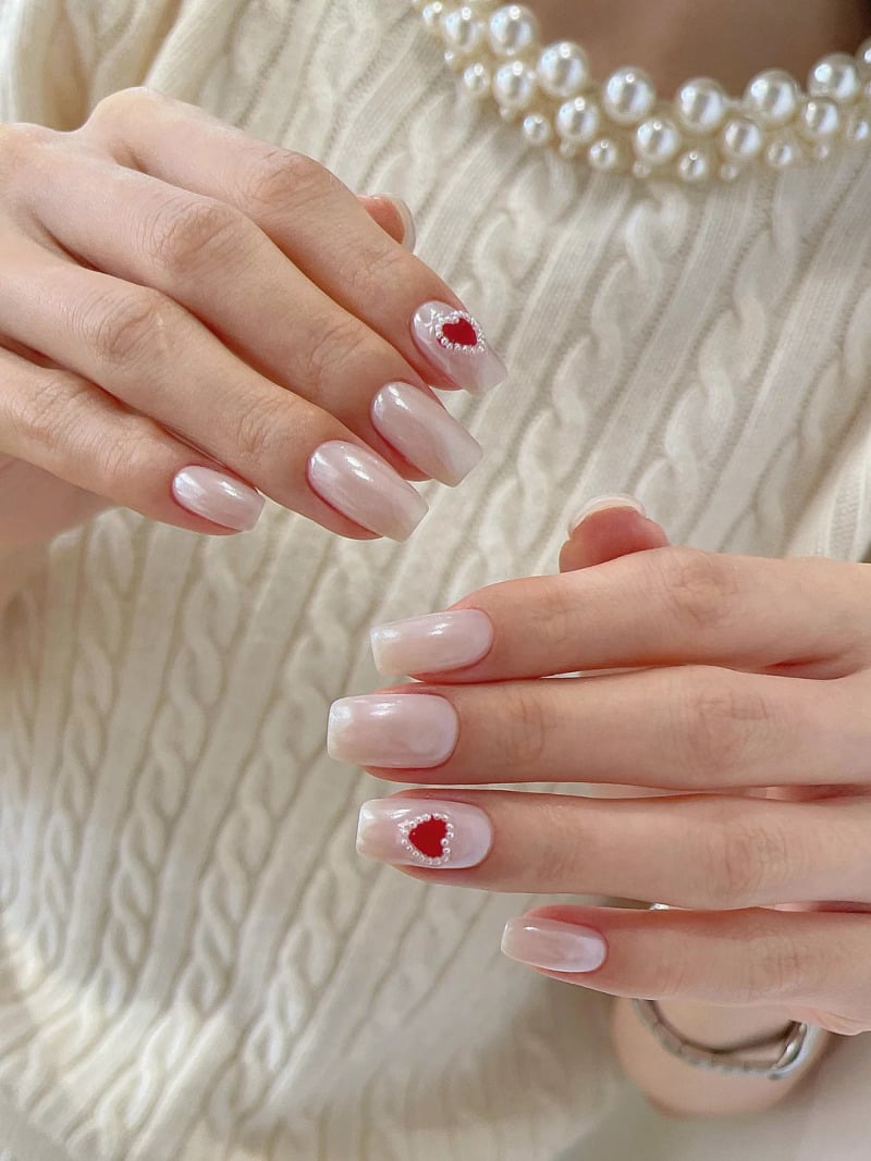 mau-nail-do-trang-cardina-4-jpg_eb7163c610c447aba9cdca229c5d6a66 Bàn tay với móng tay sơn màu nhạt, một móng trang trí hình trái tim đỏ, nền áo len trắng và dây chuyền ngọc.