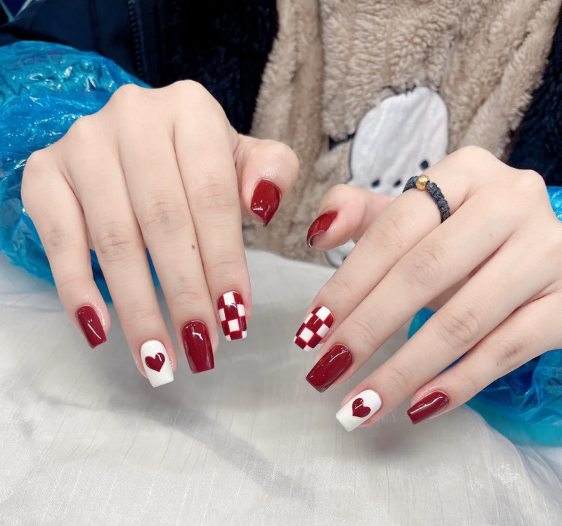 mau-nail-do-trang-cardina-6-jpg_eb2bc57957b0407cb10b24b5232fbc3d Bàn tay với móng tay được sơn màu đỏ và trắng, có họa tiết trái tim và ô vuông.
