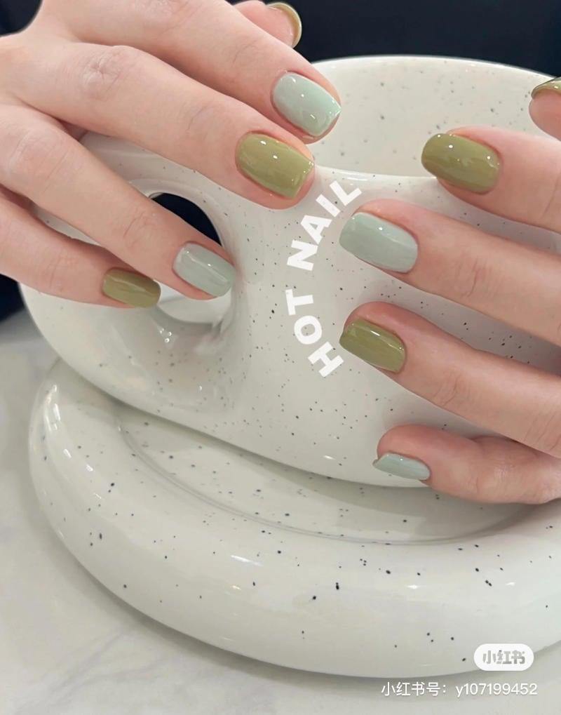 mau-nail-don-gian-cardina__111__8de21b8cd72a4dd0aa9519652485dc2d Bàn tay với móng tay sơn màu xanh lá và xám đang cầm một chiếc ly tròn màu trắng có chấm đen.