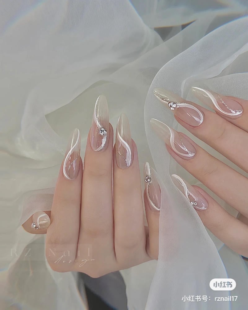 mau-nail-don-gian-cardina__114__cd85188f11e2417eb97d5f03df181831 Bàn tay với móng tay dài, sơn bóng, trang trí họa tiết và đính đá, trên nền vải trắng mềm mại.