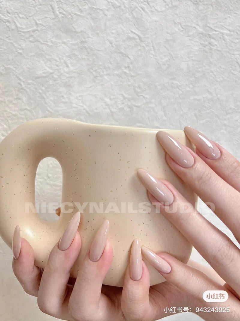 mau-nail-don-gian-cardina__124__87b10ed3f82a413ebb5fe85ccb5f35e8 Bàn tay với móng tay dài, màu nude đang cầm cốc sứ màu be trên nền tường trắng.