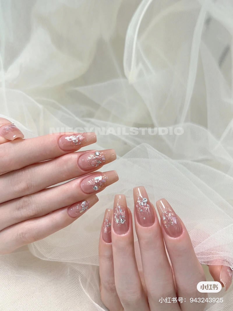 mau-nail-don-gian-cardina__127__5a481ff20d6a4450a9789932ec2c975a Bàn tay với móng tay màu hồng nhạt, trang trí hoa văn ánh bạc, trên nền vải trắng mờ.