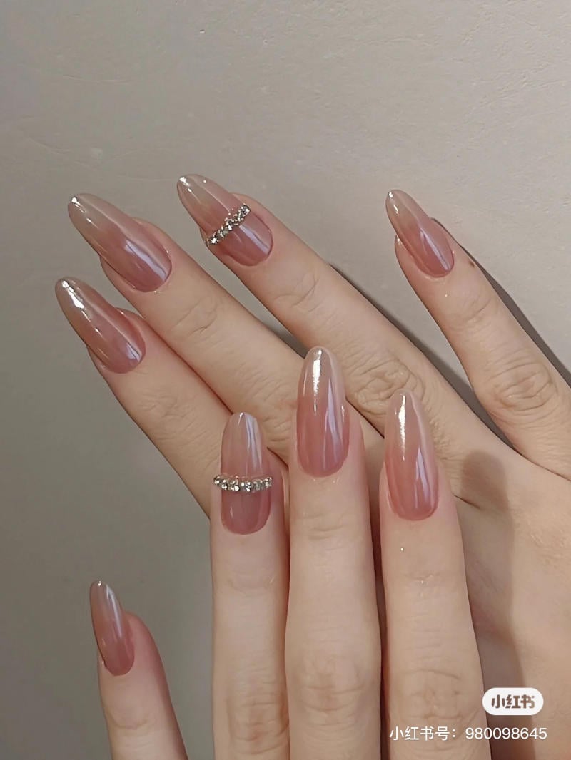 mau-nail-don-gian-cardina__138__be697df071bc4b15ab166b6babb2bccd Bàn tay với móng tay dài, màu nude bóng, có trang trí đá lấp lánh trên một số móng.