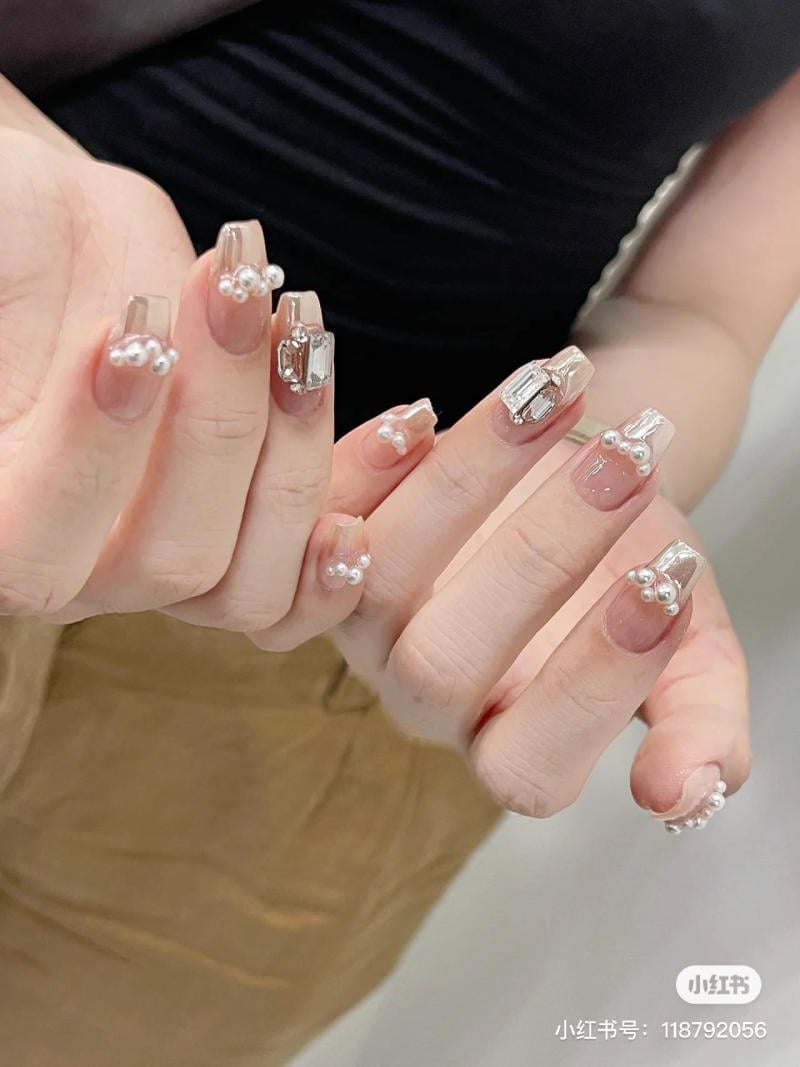 mau-nail-don-gian-cardina__139__fc4061c301cd4c679287616bf32aedc1 Bàn tay với móng tay được trang trí bằng đá và ngọc trai nhỏ, nền màu nâu nhạt.