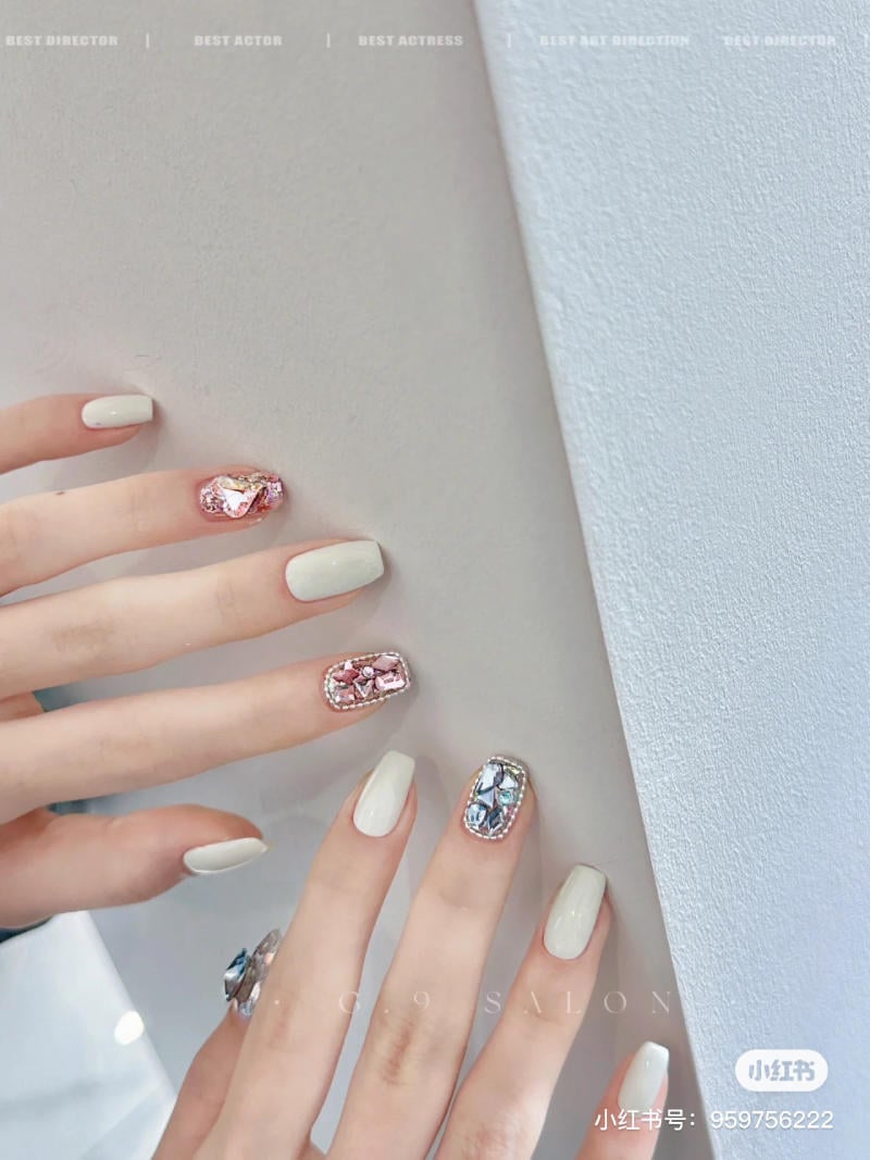 mau-nail-don-gian-cardina__142__c337490d0b17409e9d223316c7e8c33b Bàn tay với móng tay sơn màu trắng và trang trí đá quý lấp lánh trên hai ngón, đặt trên nền sáng.