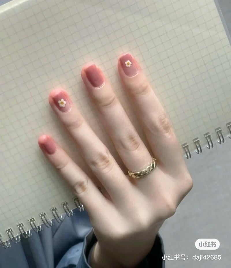 mau-nail-don-gian-cardina__147__b20afa7f939e4a92ab1f4a2fe5f5c4f1 Một bàn tay đeo nhẫn vàng, móng tay sơn màu hồng với họa tiết hoa nhỏ, cầm quyển sổ kẻ ô vuông.