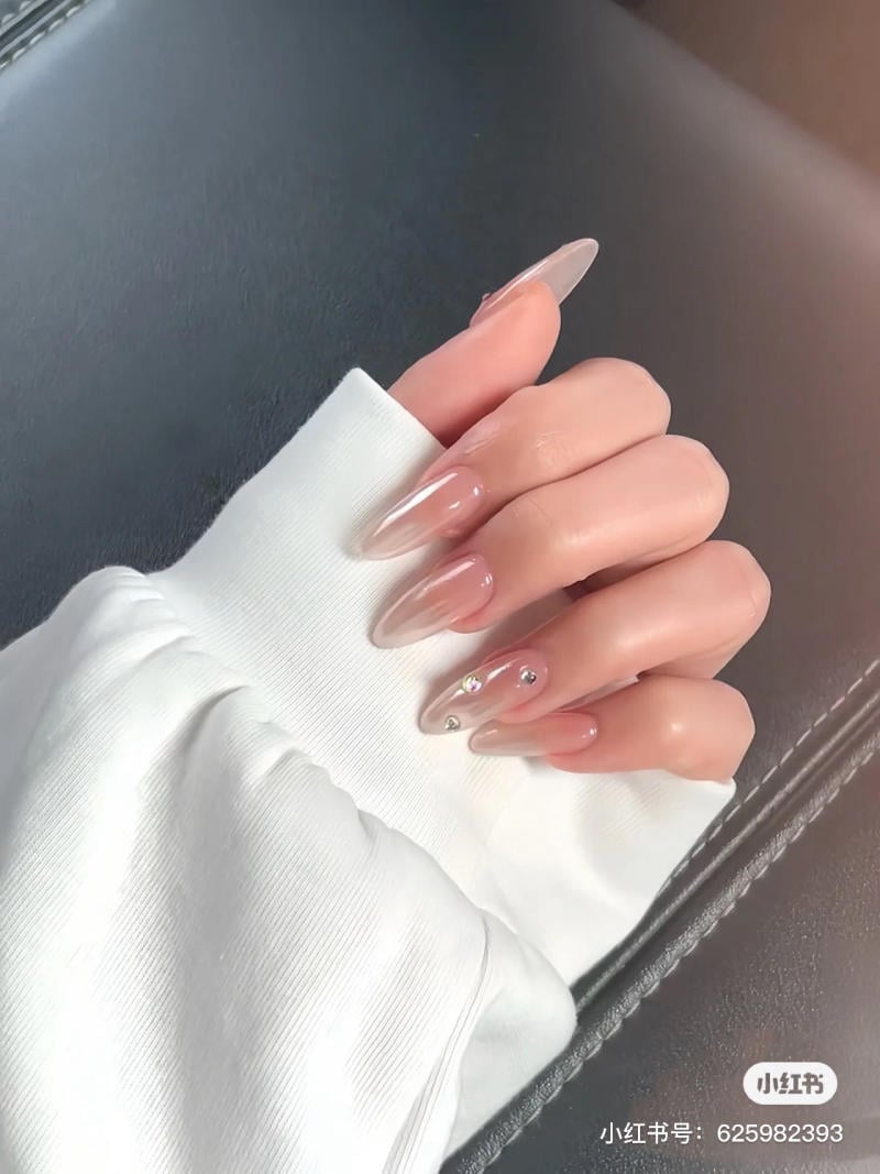 mau-nail-don-gian-cardina__151__3d3ee95e47be41339cab3627a0d60294 Bàn tay với móng tay dài, sơn màu nude, có đính đá, đặt trên nền vải màu trắng.