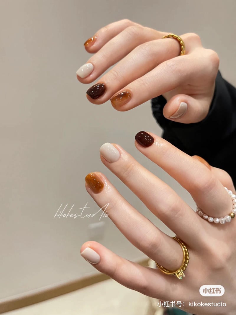 mau-nail-don-gian-cardina__20__8da2842326694271a25c9cb0b1d84f59 Bàn tay với móng tay được sơn màu nâu và kem, đeo nhẫn vàng và ngọc trai.