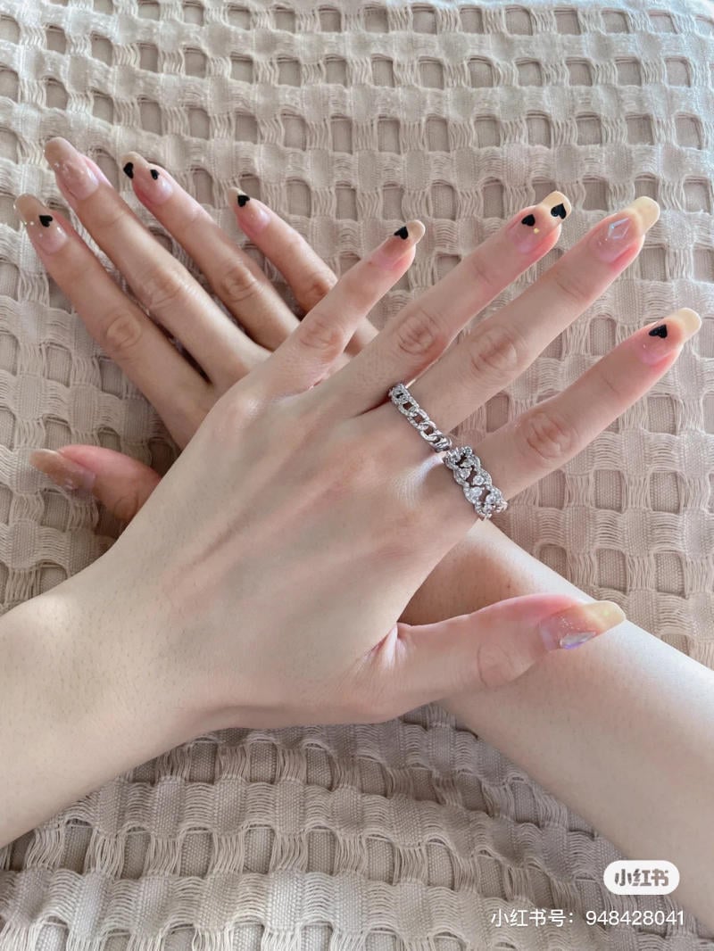 mau-nail-don-gian-cardina__94__4f1b09ebd21c474ab17b1c651c370025 Hai bàn tay đeo nhẫn, móng tay trang trí trái tim đen, đặt trên bề mặt vải màu be.