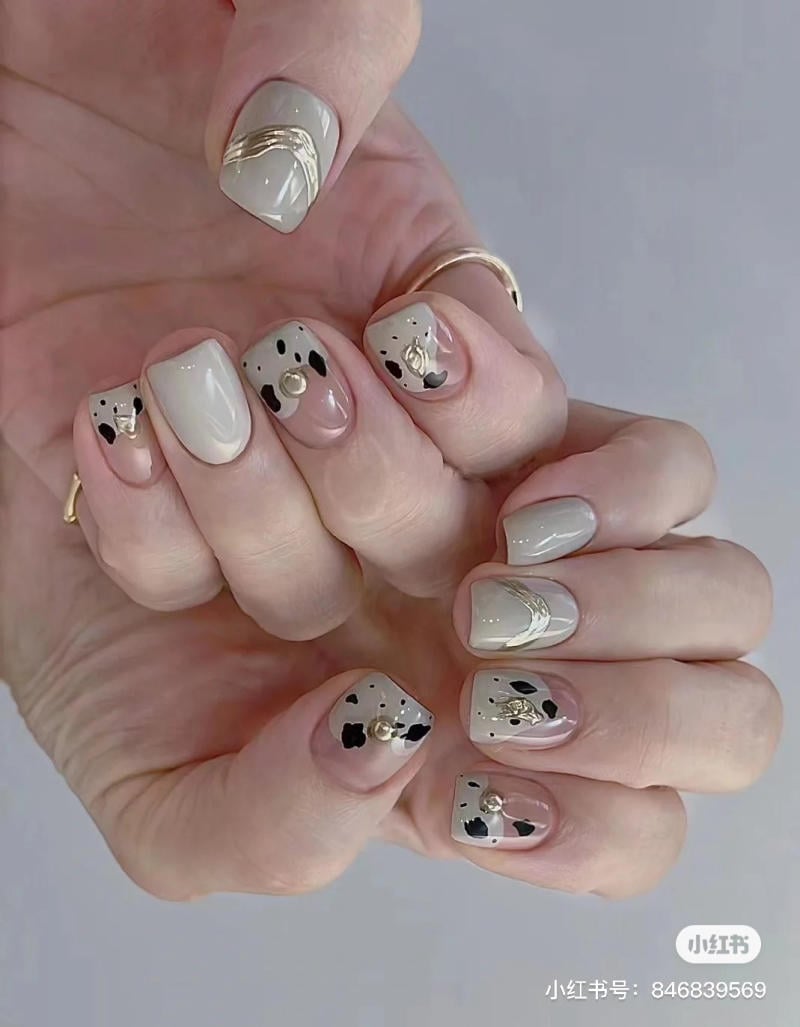 mau-nail-don-gian-cardina__97__55ce309058254e9fb753fd3ec1727731 Bàn tay với móng tay trang trí họa tiết chó đốm và phụ kiện vàng trên nền màu xám nhạt và hồng nhạt.