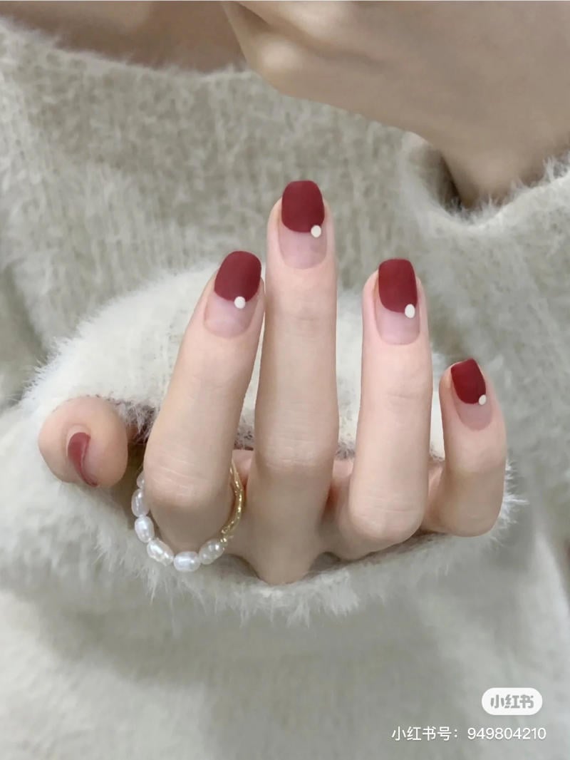 Bàn tay với móng tay sơn màu đỏ mờ, điểm nhấn là chấm trắng, đeo nhẫn ngọc trai và áo len xám.