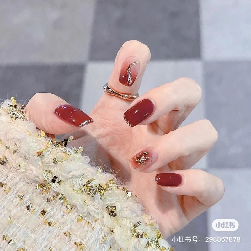 Bàn tay với móng tay sơn màu đỏ đậm, trang trí kim tuyến, nền gạch xám.
