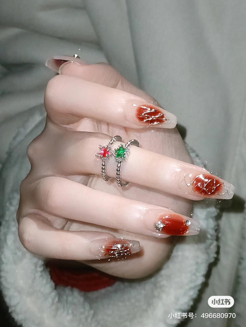 Bàn tay với móng màu đỏ đậm, trang trí họa tiết và đính đá, đeo nhẫn bạc có hạt xanh đỏ.