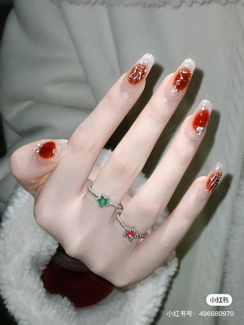 Bàn tay với móng sơn họa tiết đỏ và bạc, đeo nhẫn có đá xanh và đỏ.