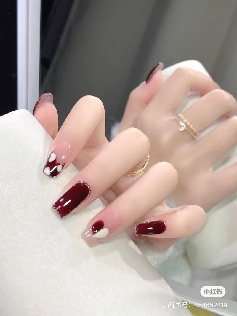 Bàn tay với móng tay được sơn màu đỏ đậm và họa tiết trắng, đeo nhẫn vàng, trên nền gương phản chiếu.