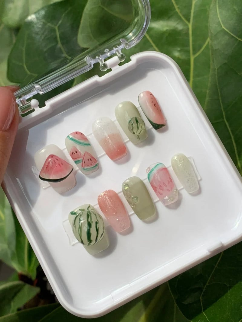 Bộ móng giả được trang trí họa tiết dưa hấu trong hộp nhựa trắng, nền lá xanh.