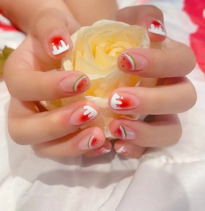 Bàn tay với móng tay vẽ hình dưa hấu, cầm một bông hoa hồng màu vàng, trên nền vải trắng và đỏ.