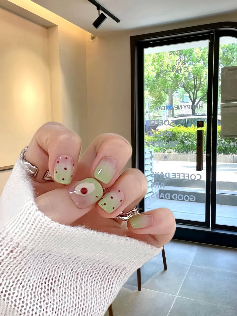 Bàn tay với móng tay được sơn màu xanh nhạt, trang trí chấm đỏ, trong không gian quán cà phê có cửa kính lớn.