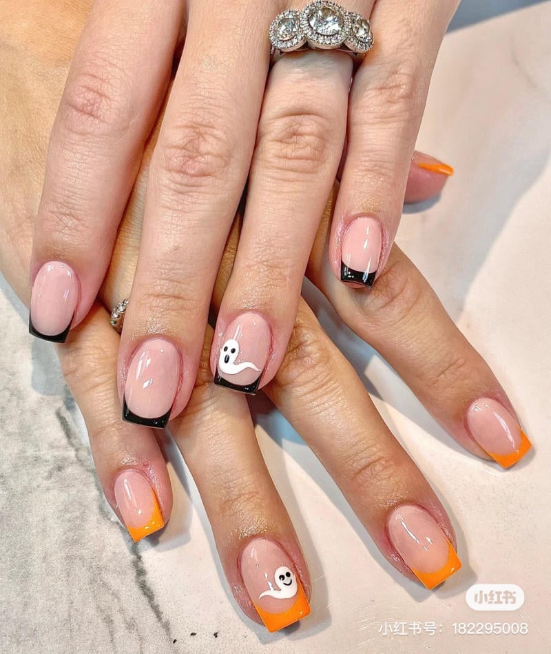 mau-nail-halloween-2023__12__36563a1c30f144baa63fa759bdb27618 Bàn tay với móng tay được sơn kiểu Pháp, trang trí hình ma nhỏ màu trắng trên nền cam và đen.