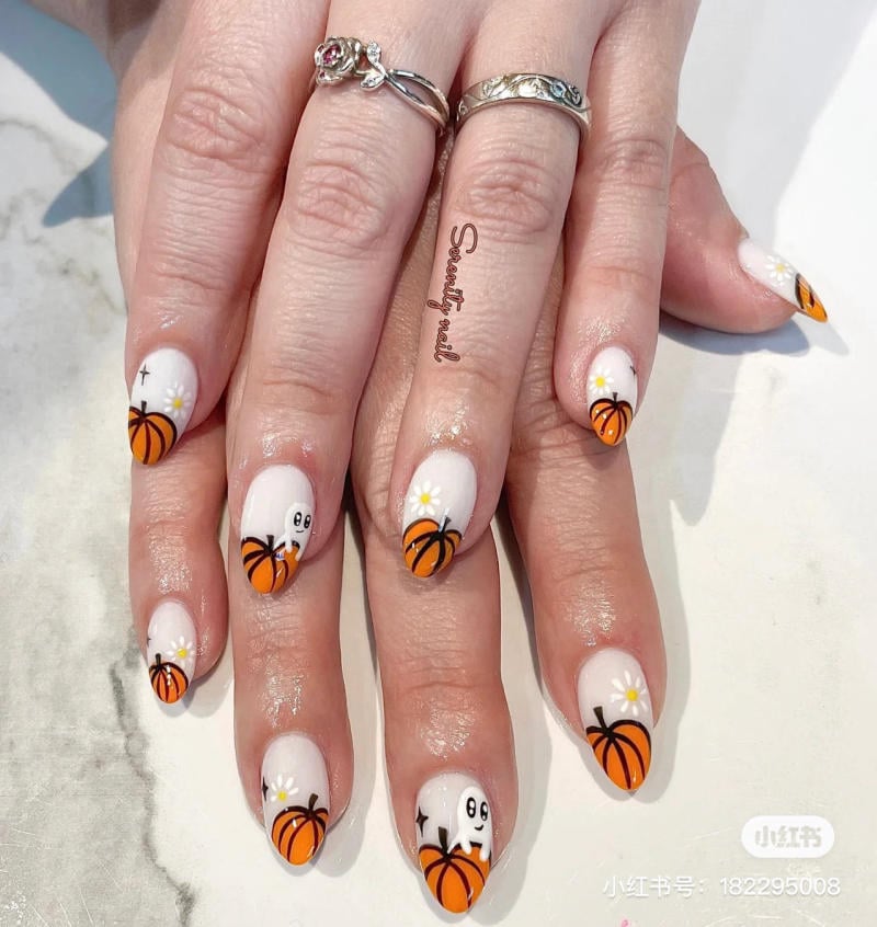mau-nail-halloween-2023__21__4be690b6f9004e9cb3be9476f182697d Bàn tay với móng tay được trang trí hình bí ngô và ma trên nền trắng, đeo nhẫn bạc có hoa văn.