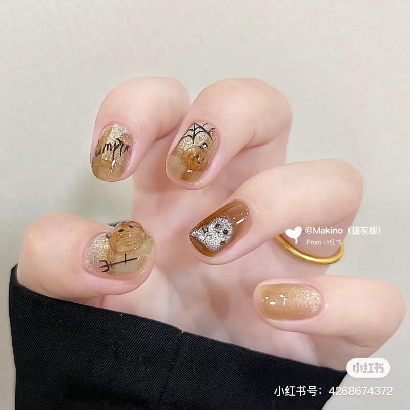 mau-nail-halloween-2023__3__ce06ed574e2d4fe4b8e3d7c29ec4722b Bàn tay với bộ móng sơn màu nâu, trang trí hình nhện và các họa tiết khác trên nền vàng lấp lánh.