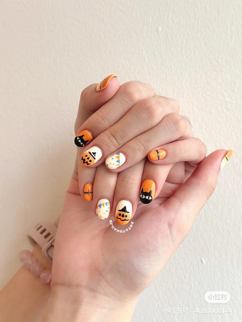 mau-nail-halloween-2023__5__eb23ce27d36441278baab71abd765f45 Bàn tay với móng tay trang trí họa tiết Halloween, gồm bí ngô, mèo đen và các biểu tượng khác trên nền cam và trắng.