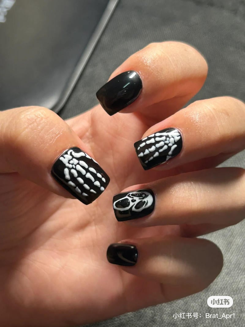 mau-nail-halloween-2023__6__a3634d92d73e474f89e037241af40e0e Bàn tay với móng tay đen, trang trí họa tiết xương trắng trên nền đen.