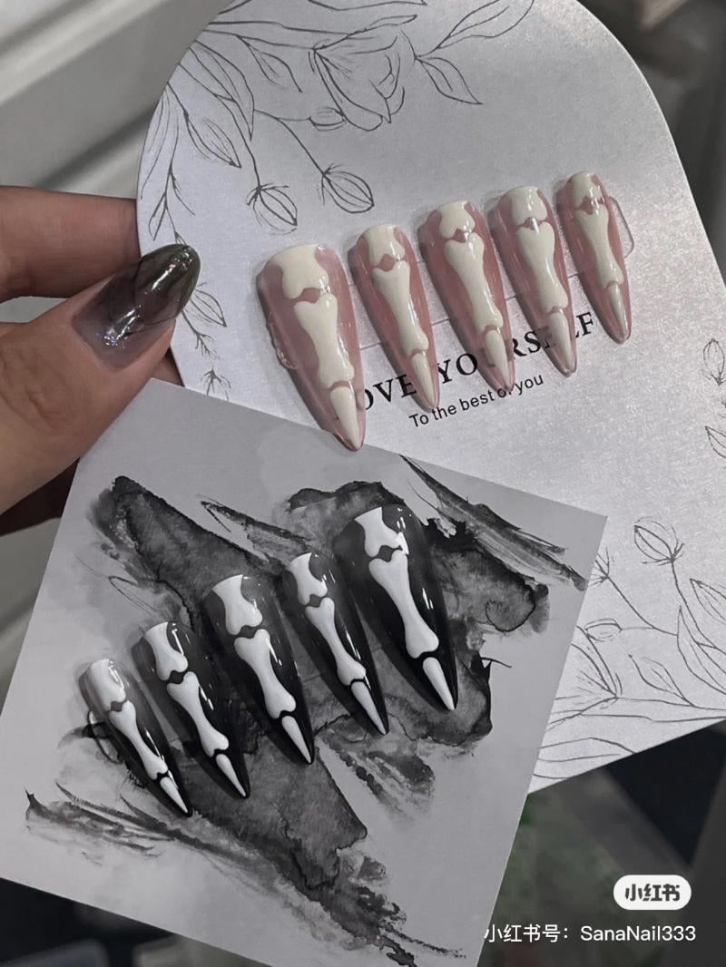 mau-nail-halloween-2023__8__73db464ef9104907b3ed6c6e483e6d80 Bộ móng giả hình xương với hai tông màu khác nhau, một màu hồng nhạt và một màu đen, trên nền trang trí hoa.