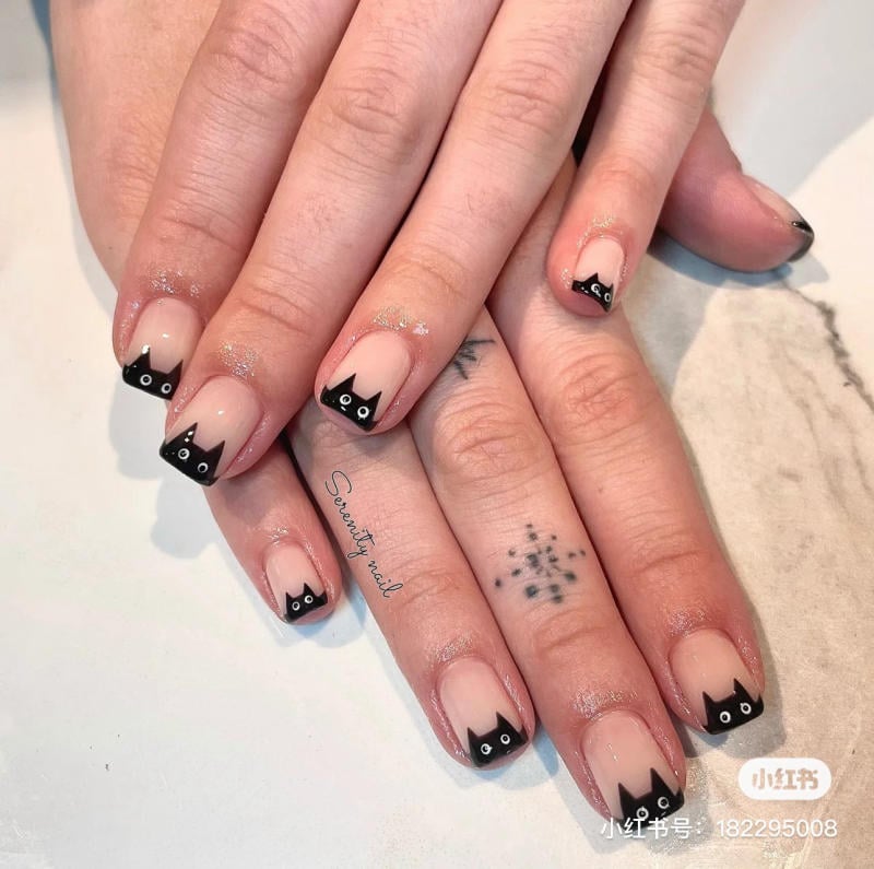 mau-nail-halloween-2023__9__5ddfeda2d1aa4791bab39a7b02466ec4 Bàn tay với móng tay được trang trí hình mèo đen trên nền sơn trong suốt, đặt trên bề mặt sáng màu.