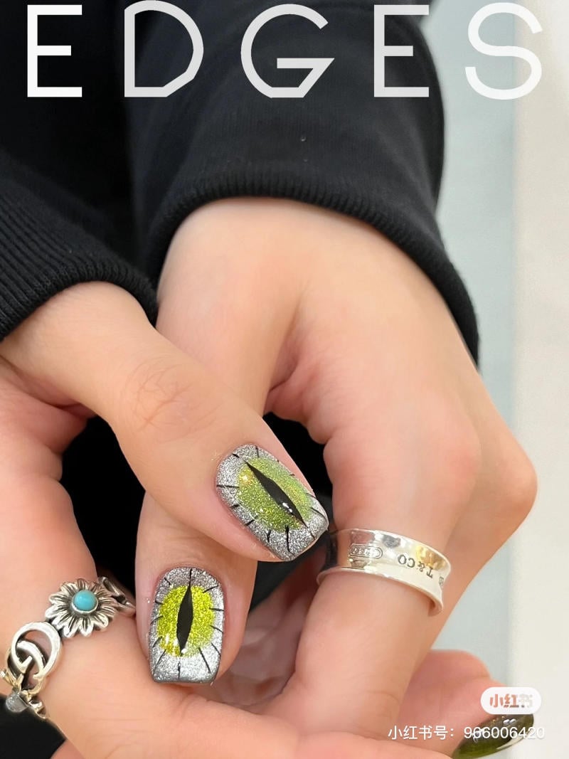 mau-nail-halloween-dep__10__6d3fc4f405614bad9fa7a6b9a153230d Bàn tay với móng tay được trang trí họa tiết mắt mèo màu xanh, đeo nhẫn bạc và áo đen.