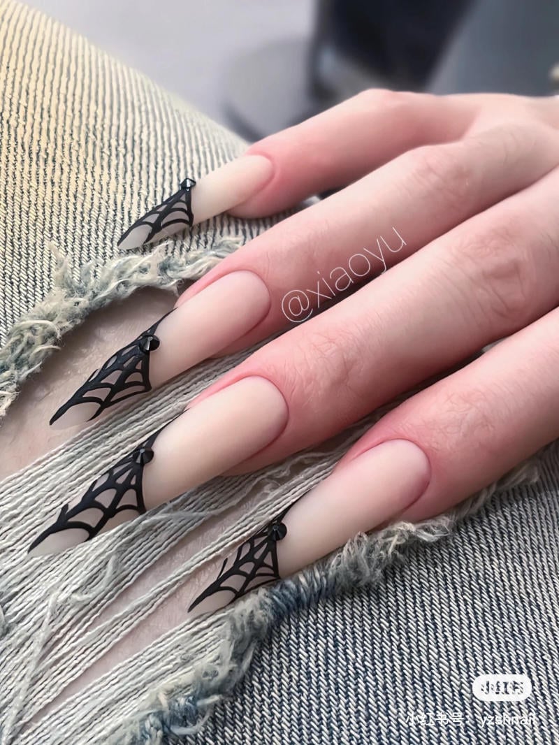 mau-nail-halloween-dep__14__e911690922f14c53860cce8f68ffb0f6 Bàn tay với móng dài sơn màu nude, trang trí hình mạng nhện đen, đặt trên nền vải jean rách.