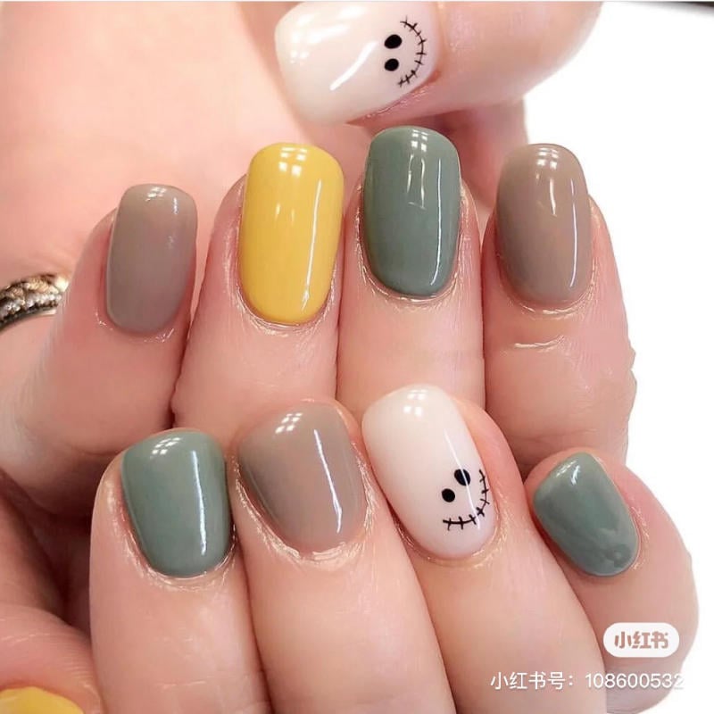 mau-nail-halloween-dep__16__50403fc8e592479dae1c7ae6bddaf1e1 Bàn tay với móng tay sơn màu xám, xanh lá, vàng và trắng có họa tiết mặt cười.